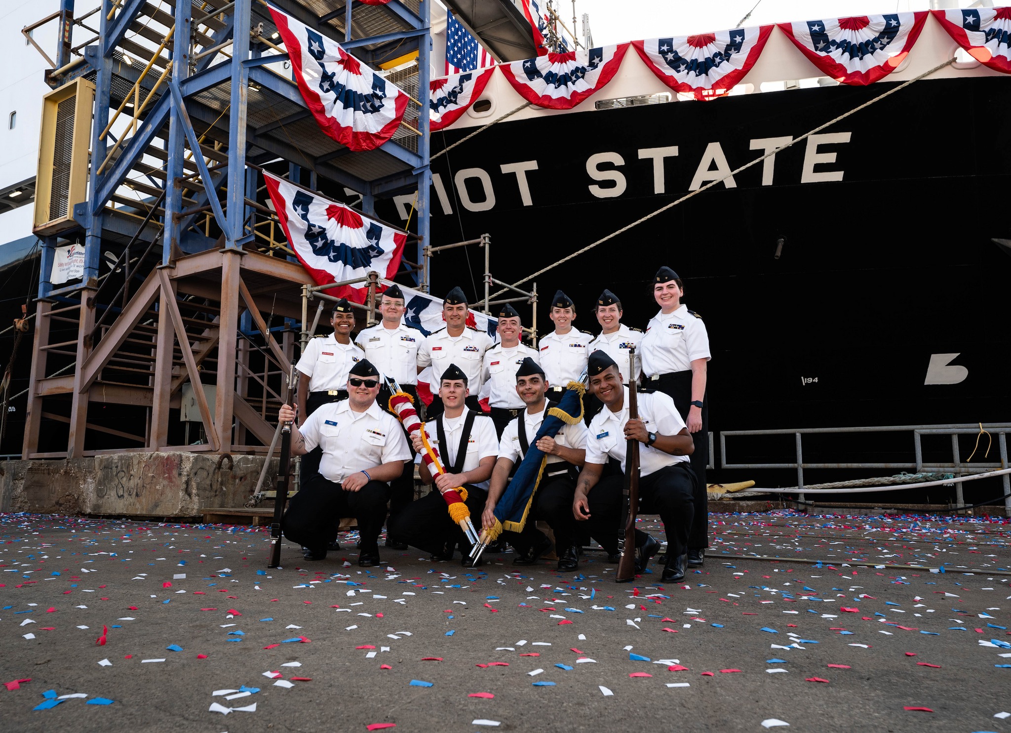 A group of 11 young men and women stand in front of a large ship covered in celebratory flags surrounded by confetti.